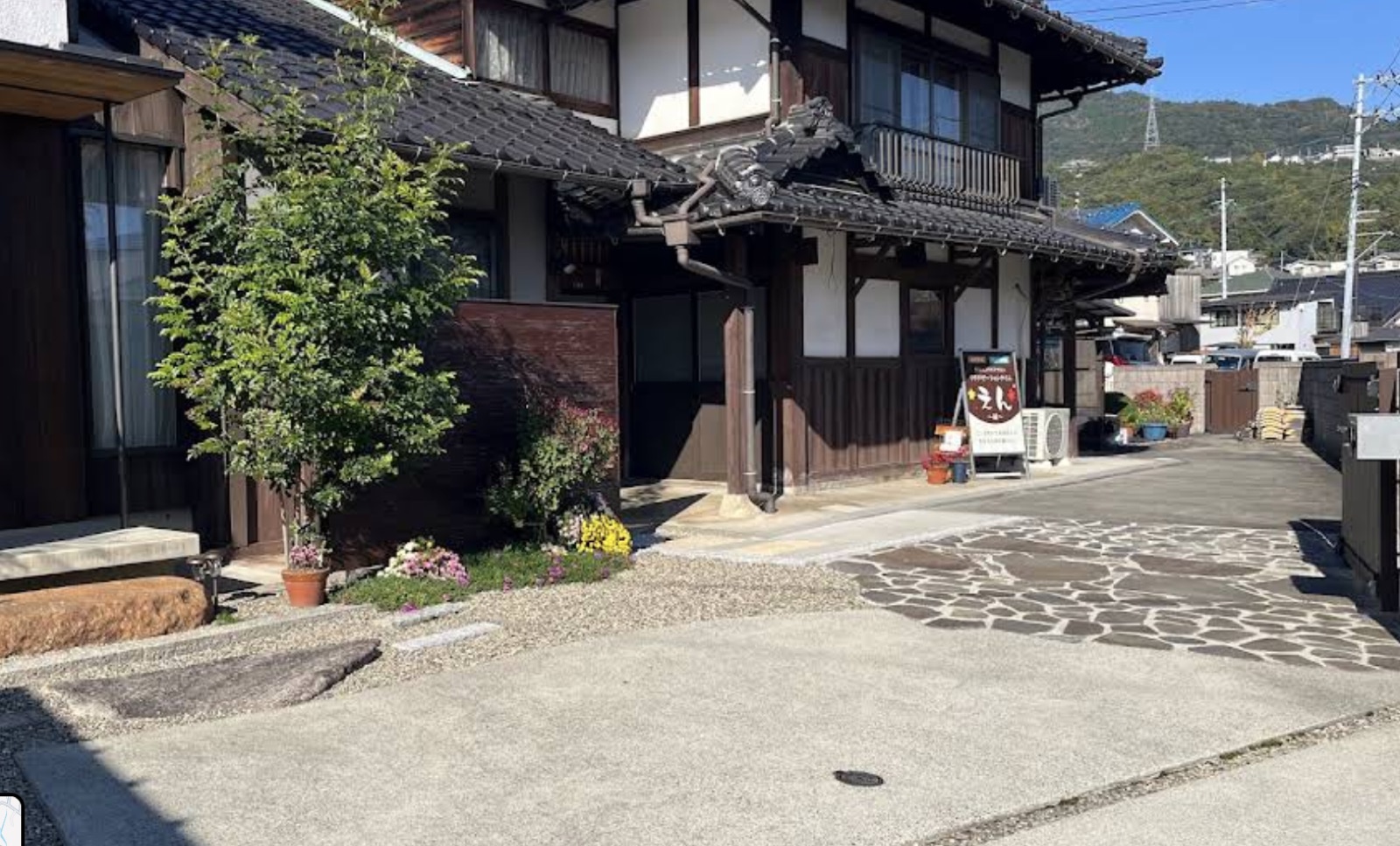 瓦屋根の日本家屋の外観。青空と背景の山に囲まれた、石畳の駐車場がある古民家サロン